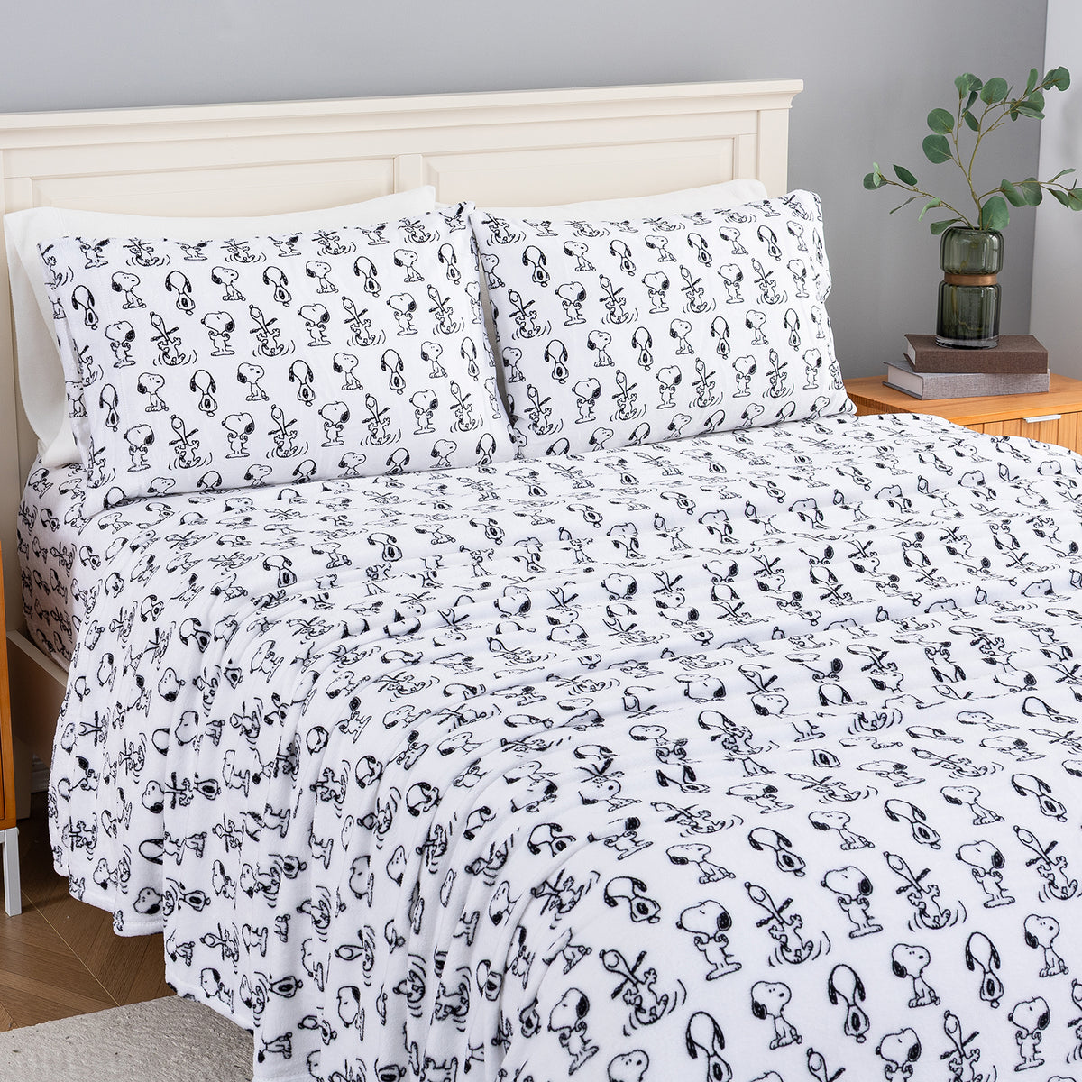A bed dressed in a white bedding set with an all-over black Snoopy character print, shown in a styled bedroom with a light wood headboard, bedside table, books, and a plant.