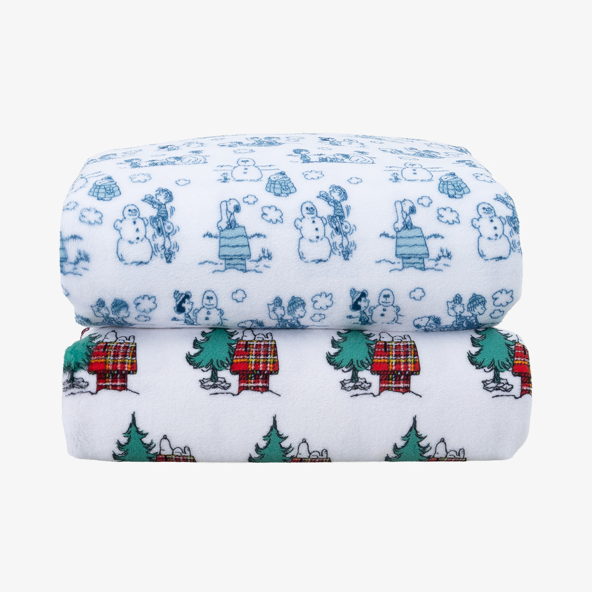 Two folded white fabric sheets with holiday cartoon prints, stacked on a light gray background. The top item has blue snowman and character illustrations, and the bottom item has green trees, red plaid doghouses, and small white figures.