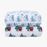 Two folded white fabric sheets with holiday cartoon prints, stacked on a light gray background. The top item has blue snowman and character illustrations, and the bottom item has green trees, red plaid doghouses, and small white figures.