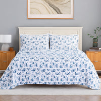 A neatly made bed in a styled bedroom setting, featuring a white comforter and matching pillow shams with a repeating blue cartoon character print. The bed is set against a light gray wall with a cream-colored wooden headboard, flanked by wooden nightstands with lamps and a small plant.