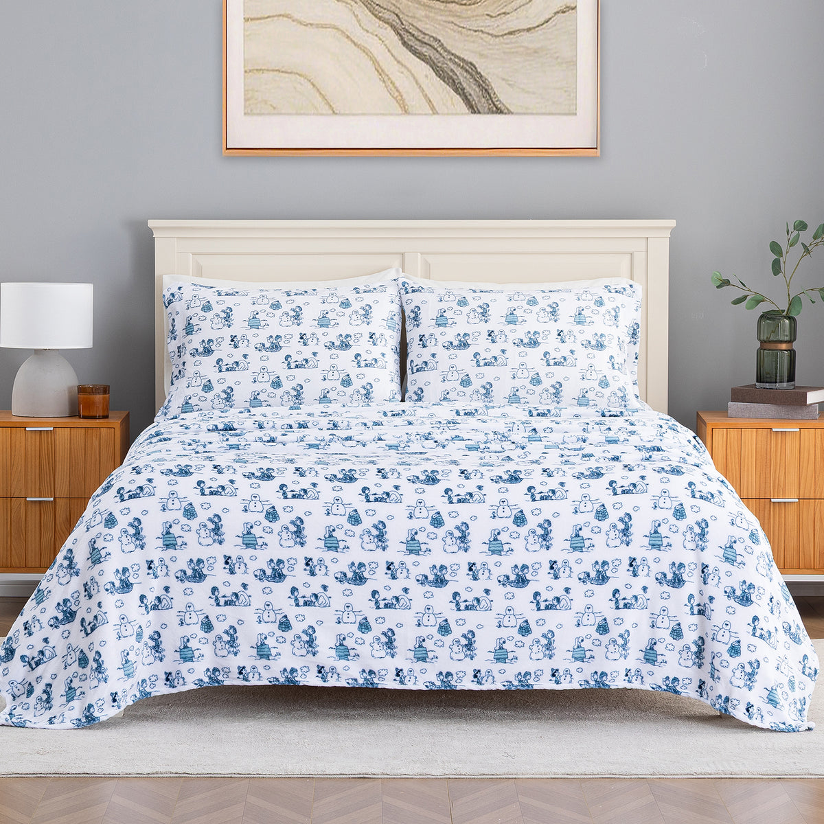 A neatly made bed in a styled bedroom setting, featuring a white comforter and matching pillow shams with a repeating blue cartoon character print. The bed is set against a light gray wall with a cream-colored wooden headboard, flanked by wooden nightstands with lamps and a small plant.