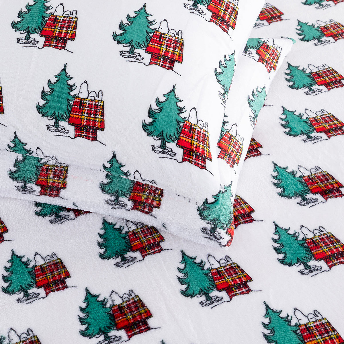 White fabric bedding set featuring a repeating print of Snoopy and Woodstock sitting on red and green plaid Christmas stockings beside green Christmas trees.
