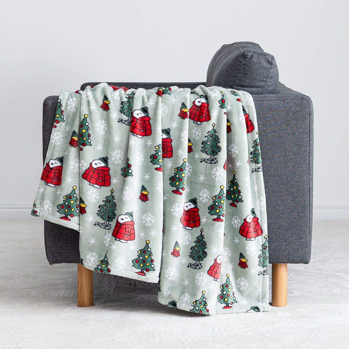 A sage green holiday Peanuts Printed VelvetLoft Throw draped over a grey chair.