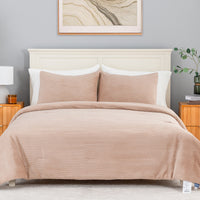 A neatly made bed in a styled bedroom setting, featuring a ribbed comforter and two matching pillow shams in a dusty blush pink color. The bed is dressed with white pillows and set against a light gray wall with a wooden headboard, flanked by wooden nightstands with a lamp and a small plant.