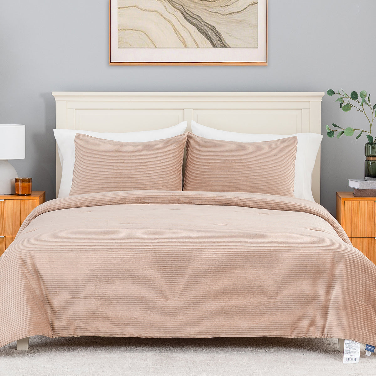 A neatly made bed in a styled bedroom setting, featuring a ribbed comforter and two matching pillow shams in a dusty blush pink color. The bed is dressed with white pillows and set against a light gray wall with a wooden headboard, flanked by wooden nightstands with a lamp and a small plant.