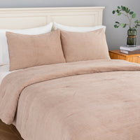 A bed dressed in a warm beige corduroy comforter and two matching pillowcases, styled in a bedroom with a cream headboard, wooden nightstand, books, and a green glass vase with leaves.
