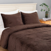 A bed dressed in a dark brown corduroy comforter  and matching pillow shams, styled in a bedroom with a white headboard, white pillows, a wooden nightstand, books, and a green glass vase with a plant.