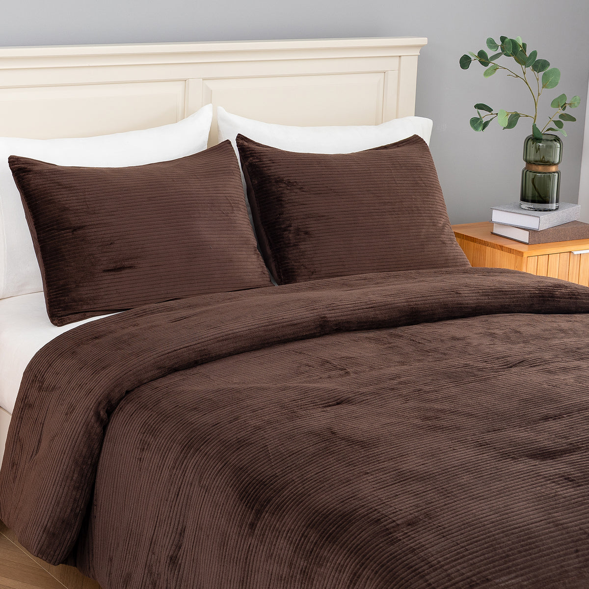 A bed dressed in a dark brown corduroy comforter  and matching pillow shams, styled in a bedroom with a white headboard, white pillows, a wooden nightstand, books, and a green glass vase with a plant.