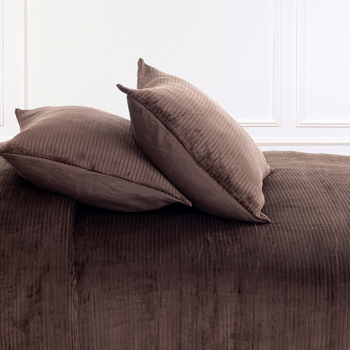 Two brown corduroy pillows resting on a matching brown corduroy bedspread or sofa, set against a white paneled wall.