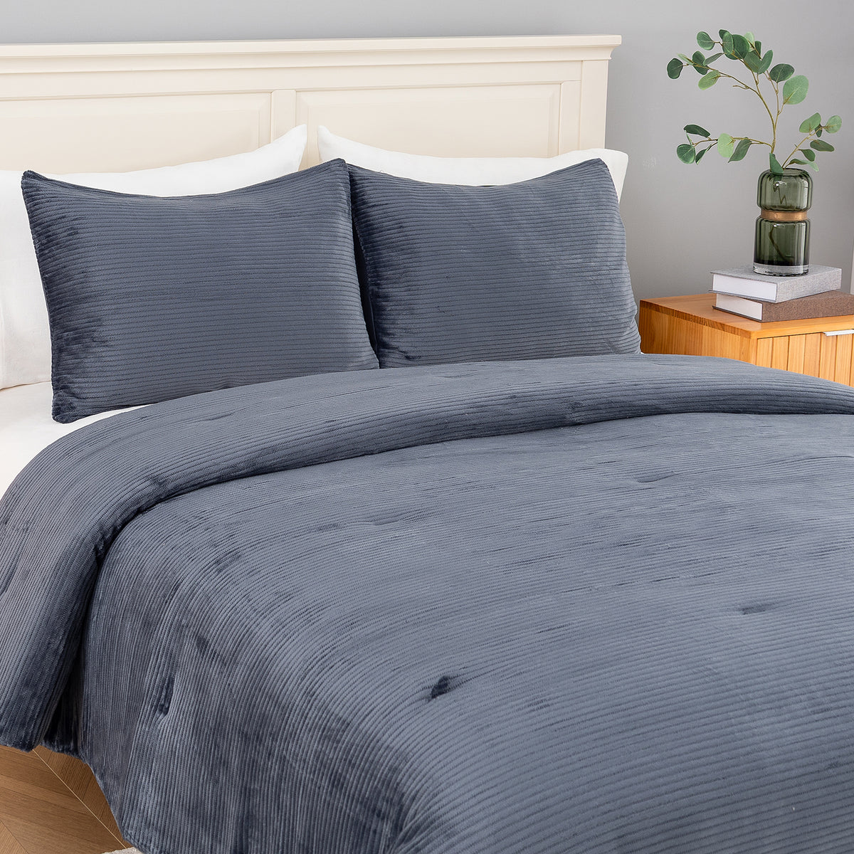 A bed dressed in a slate blue corduroy comforter and matching pillow shams, styled in a bedroom with a white headboard, wooden nightstand, and a green glass vase with eucalyptus.