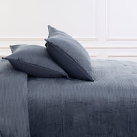 A close-up of a bed dressed in a dark slate blue corduroy bedding set, featuring a textured corduroy duvet cover and two matching pillows, set against a white paneled wall.