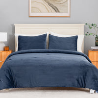 A neatly made bed in a bedroom setting, featuring a textured navy blue corduroy or ribbed comforter and matching pillow shams, set against a light gray wall with a cream headboard and wooden nightstands.