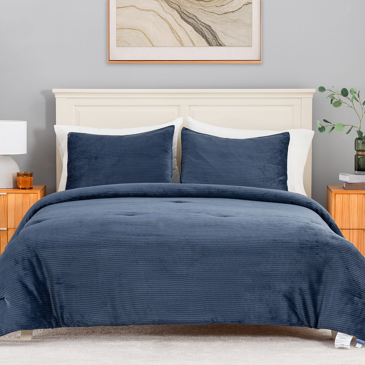 A neatly made bed in a bedroom setting, featuring a textured navy blue corduroy or ribbed comforter and matching pillow shams, set against a light gray wall with a cream headboard and wooden nightstands.