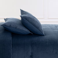 A close-up of a blue corduroy upholstered bed or sofa with two matching blue pillows against a white paneled wall.