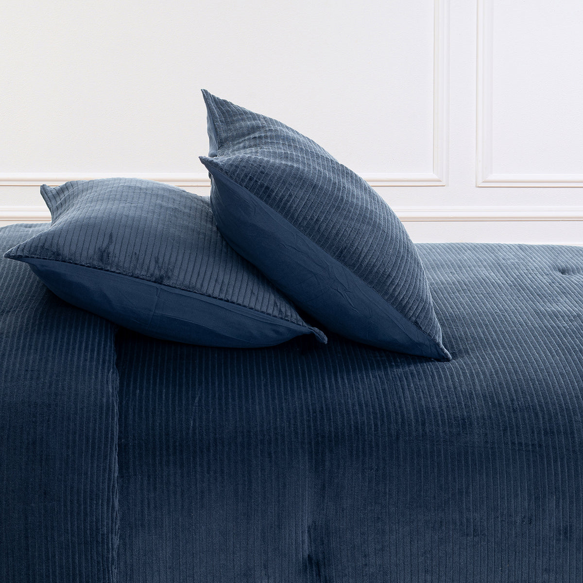 A close-up of a blue corduroy upholstered bed or sofa with two matching blue pillows against a white paneled wall.