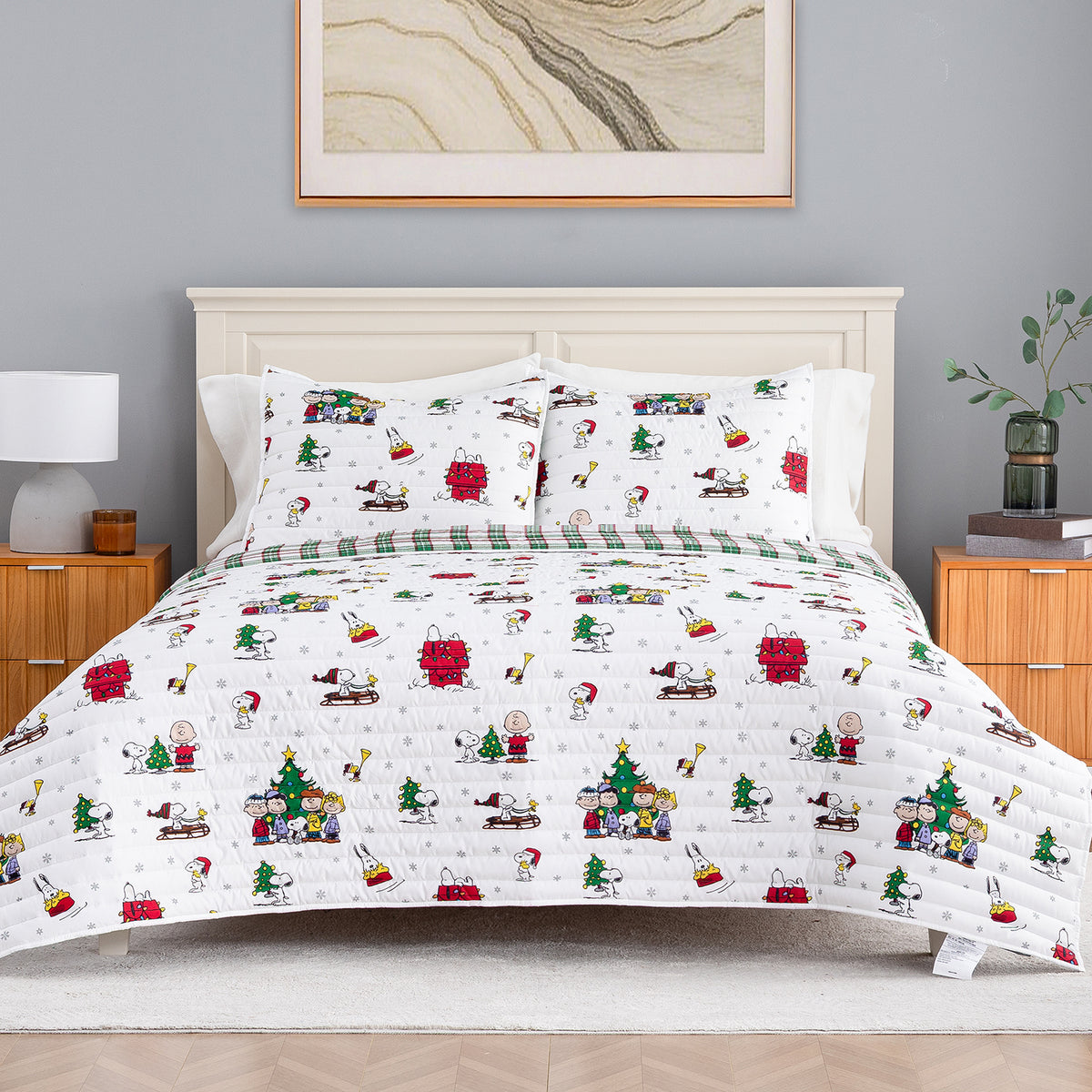 A festive holiday-themed bedding set featuring a white quilt and matching pillow shams printed with cartoon characters like Snoopy and Woodstock in Christmas scenes, styled on a bed in a modern bedroom setting.