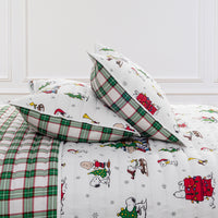 A festive holiday-themed bedding set featuring a white quilted cover with Snoopy and Woodstock Christmas prints, matching pillows, and green and red plaid accents, styled in a bedroom setting.