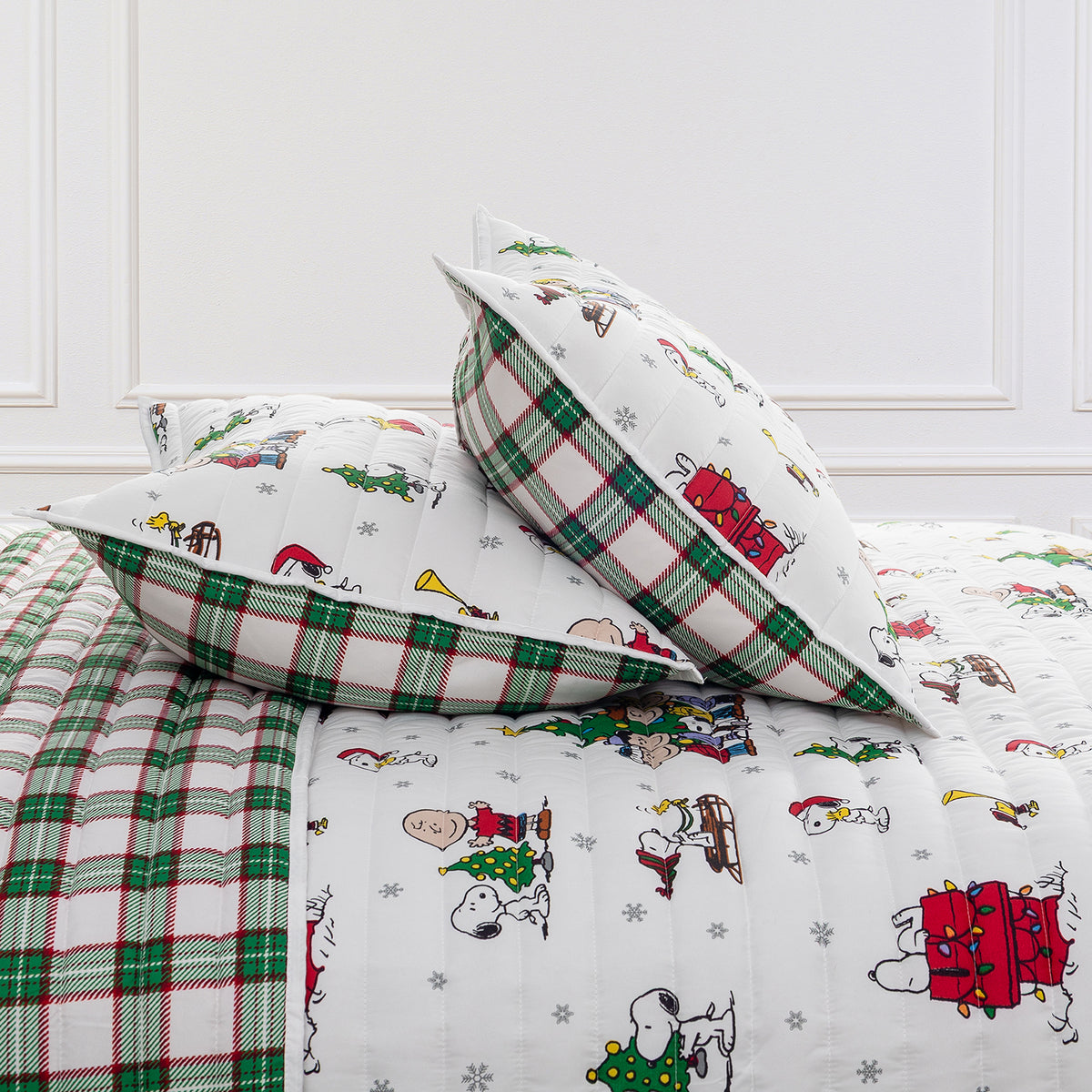 A festive holiday-themed bedding set featuring a white quilted cover with Snoopy and Woodstock Christmas prints, matching pillows, and green and red plaid accents, styled in a bedroom setting.