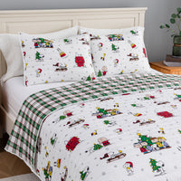A holiday-themed bedding set featuring a white comforter and matching pillowcases with a colorful cartoon print of Snoopy, Woodstock, Charlie Brown, and other Peanuts characters, alongside a green and white plaid blanket section, styled on a bed with a cream headboard.