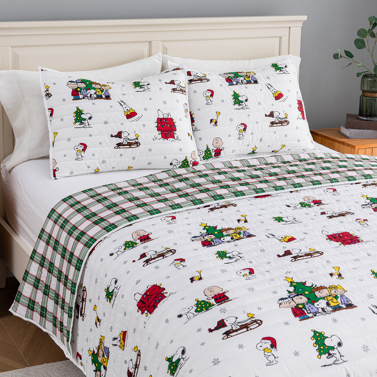 A holiday-themed bedding set featuring a white comforter and matching pillowcases with a colorful cartoon print of Snoopy, Woodstock, Charlie Brown, and other Peanuts characters, alongside a green and white plaid blanket section, styled on a bed with a cream headboard.