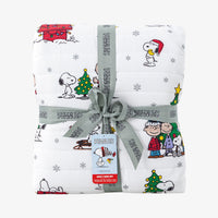 A folded white quilted blanket featuring a colorful Peanuts holiday print, wrapped with a gray ribbon and bow. The blanket displays characters like Snoopy, Woodstock, Charlie Brown, and Peanuts.