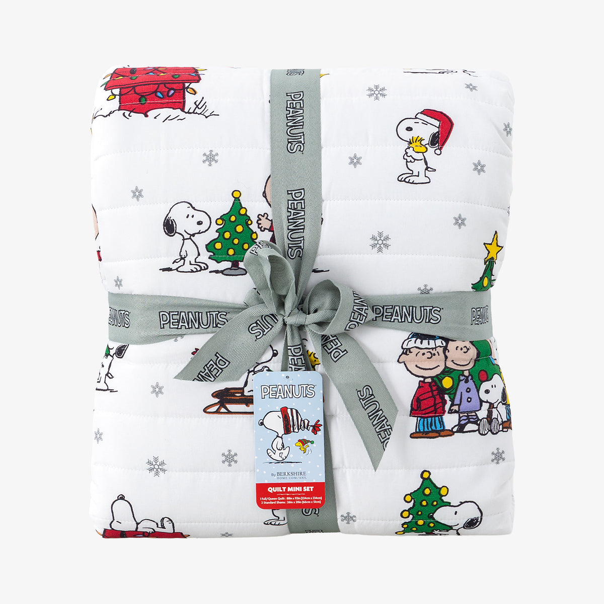 A folded white quilted blanket featuring a colorful Peanuts holiday print, wrapped with a gray ribbon and bow. The blanket displays characters like Snoopy, Woodstock, Charlie Brown, and Peanuts.