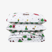 A stack of three folded white quilted pillows and a matching folded comforter set, printed with a festive holiday cartoon pattern featuring Snoopy, Peanuts characters, Christmas trees, snowflakes, and green plaid borders, on a light gray background.