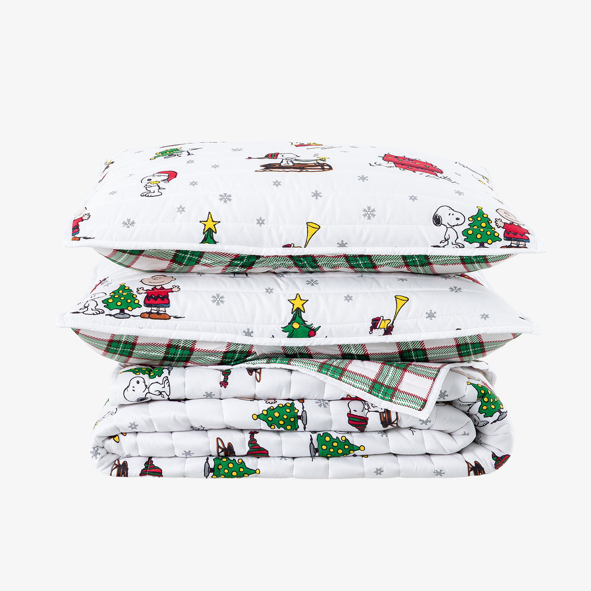 A stack of three folded white quilted pillows and a matching folded comforter set, printed with a festive holiday cartoon pattern featuring Snoopy, Peanuts characters, Christmas trees, snowflakes, and green plaid borders, on a light gray background.