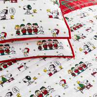 A close-up of a quilted bedding set featuring the Peanuts characters Snoopy, Woodstock, Charlie Brown, and the other Peanuts gang in Christmas outfits, with red piping and a red plaid reverse side.