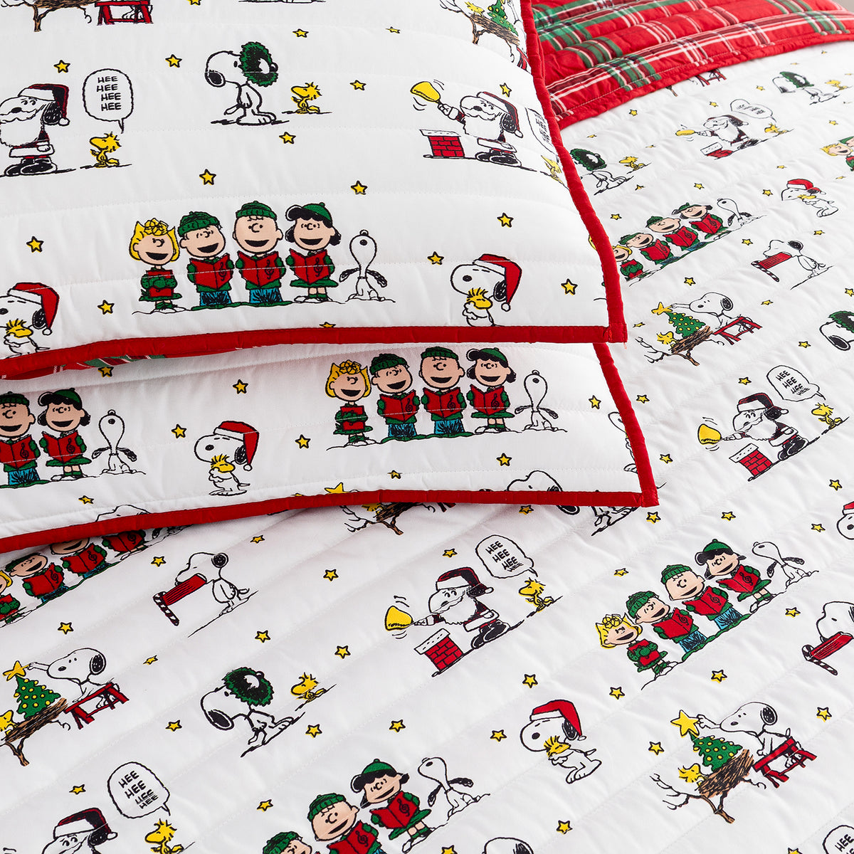 A close-up of a quilted bedding set featuring the Peanuts characters Snoopy, Woodstock, Charlie Brown, and the other Peanuts gang in Christmas outfits, with red piping and a red plaid reverse side.