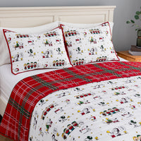 A bed dressed in a festive holiday-themed bedding set featuring a white background printed with classic cartoon characters, red trim, and a red plaid blanket.