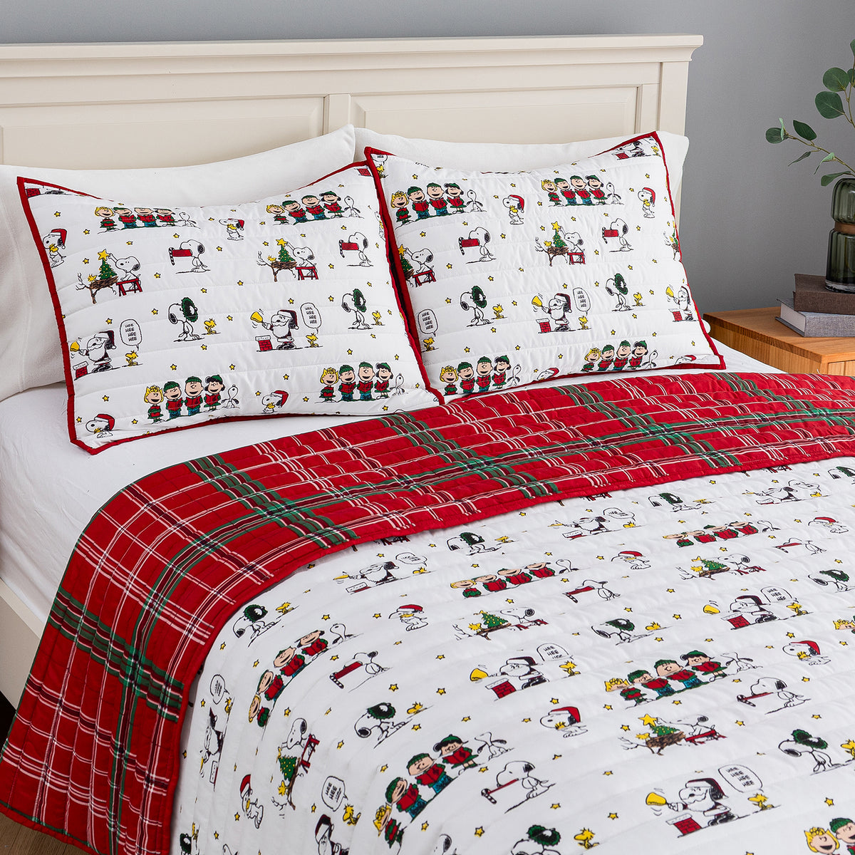 A bed dressed in a festive holiday-themed bedding set featuring a white background printed with classic cartoon characters, red trim, and a red plaid blanket.