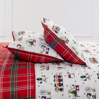 A festive bed set featuring a white quilted cover and matching pillowcases printed with classic cartoon characters like Snoopy and Woodstock, accented with red and green plaid borders and a red plaid blanket.