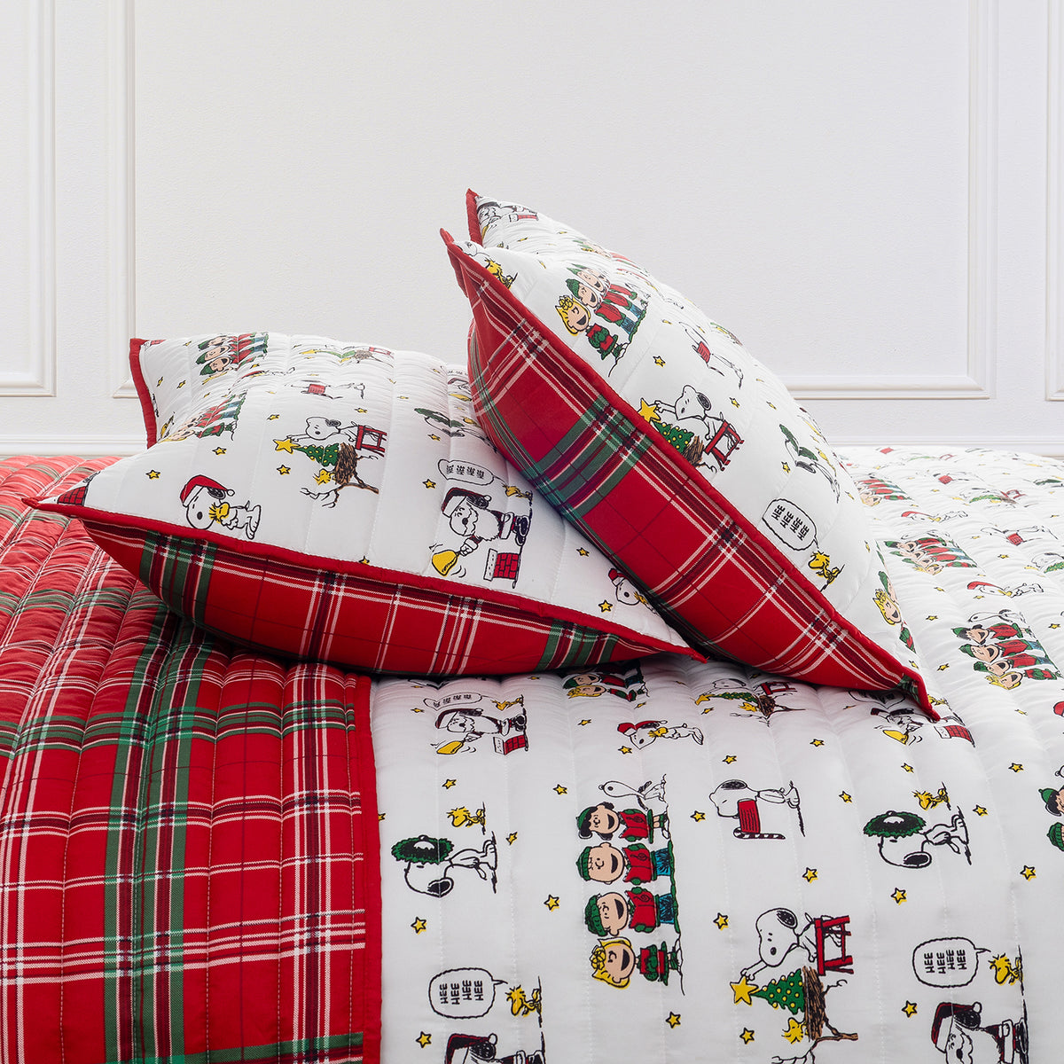 A festive bed set featuring a white quilted cover and matching pillowcases printed with classic cartoon characters like Snoopy and Woodstock, accented with red and green plaid borders and a red plaid blanket.