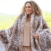A smiling woman with curly hair wearing a plush leopard-print faux fur blanket over a beige knit top, photographed outdoors with a softly blurred green background.