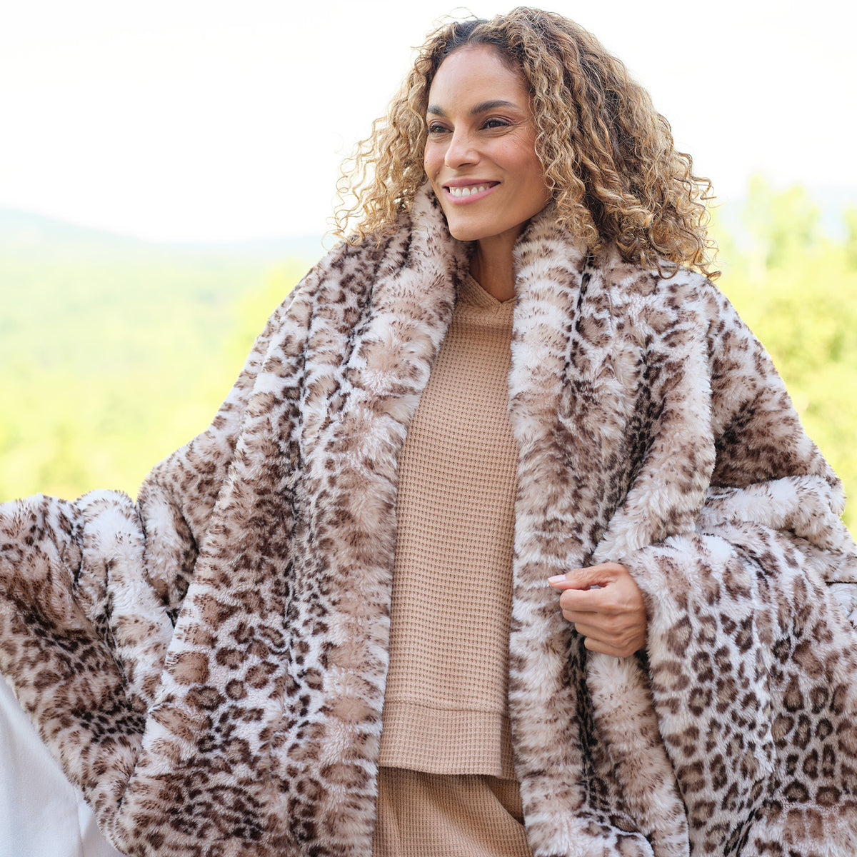 A smiling woman with curly hair wearing a plush leopard-print faux fur blanket over a beige knit top, photographed outdoors with a softly blurred green background.