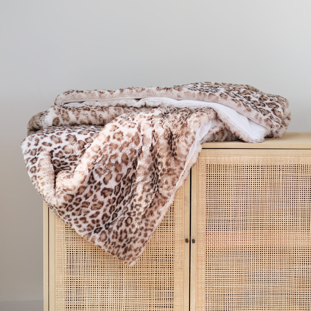 A plush faux-fur throw blanket with a brown and beige leopard print pattern and a white reverse side, draped over a light wood cabinet with a woven rattan front.