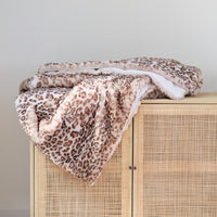 A plush faux-fur throw blanket with a brown and beige leopard print pattern and a white reverse side, draped over a light wood cabinet with a woven rattan front.