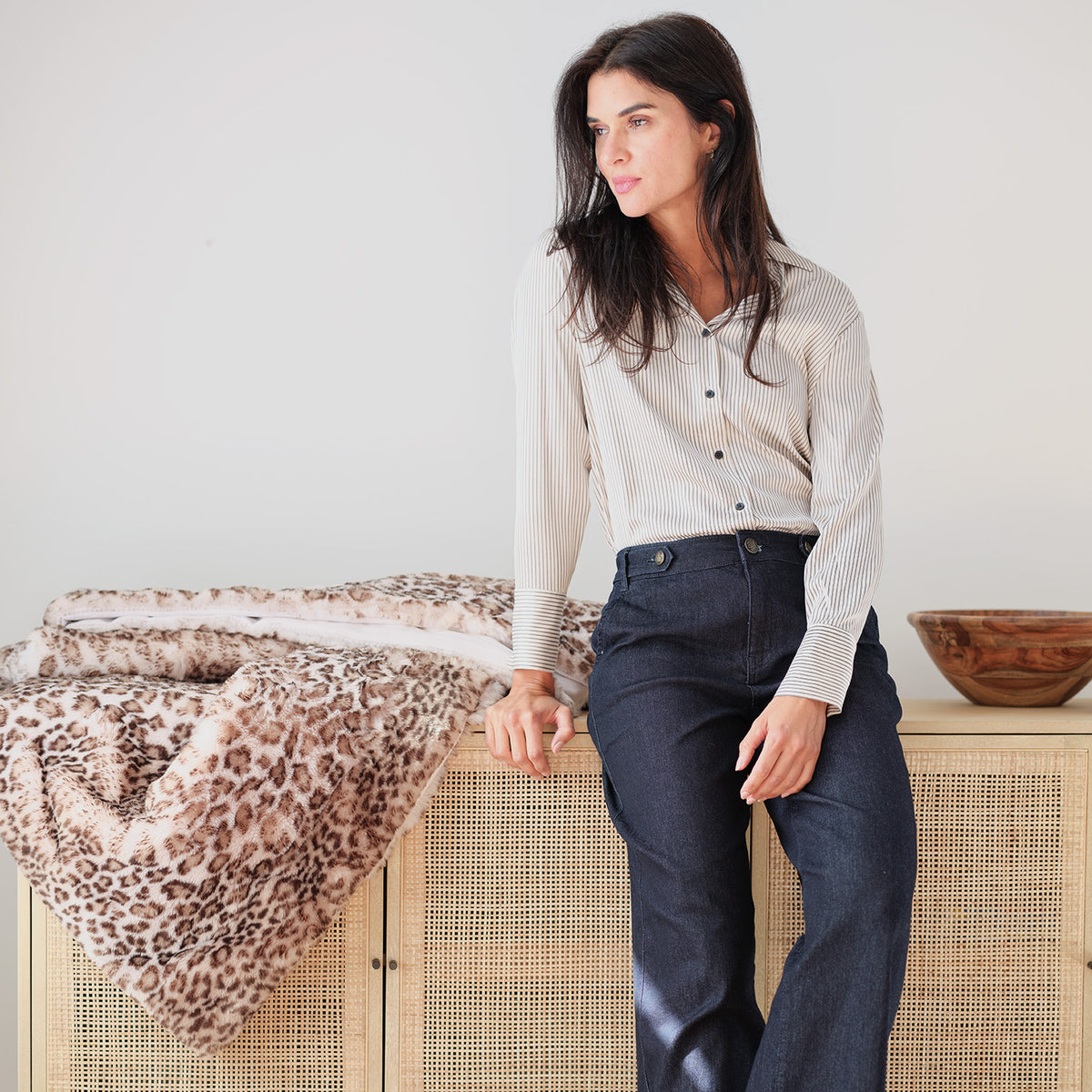 A woman wearing a cream ribbed long-sleeve button-up shirt and dark blue jeans leans against a light wood cane cabinet next to a leopard-print throw blanket.