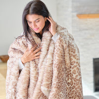 A woman wrapped in a plush faux fur throw blanket with a beige and brown leopard print pattern, standing in a brightly lit indoor setting.