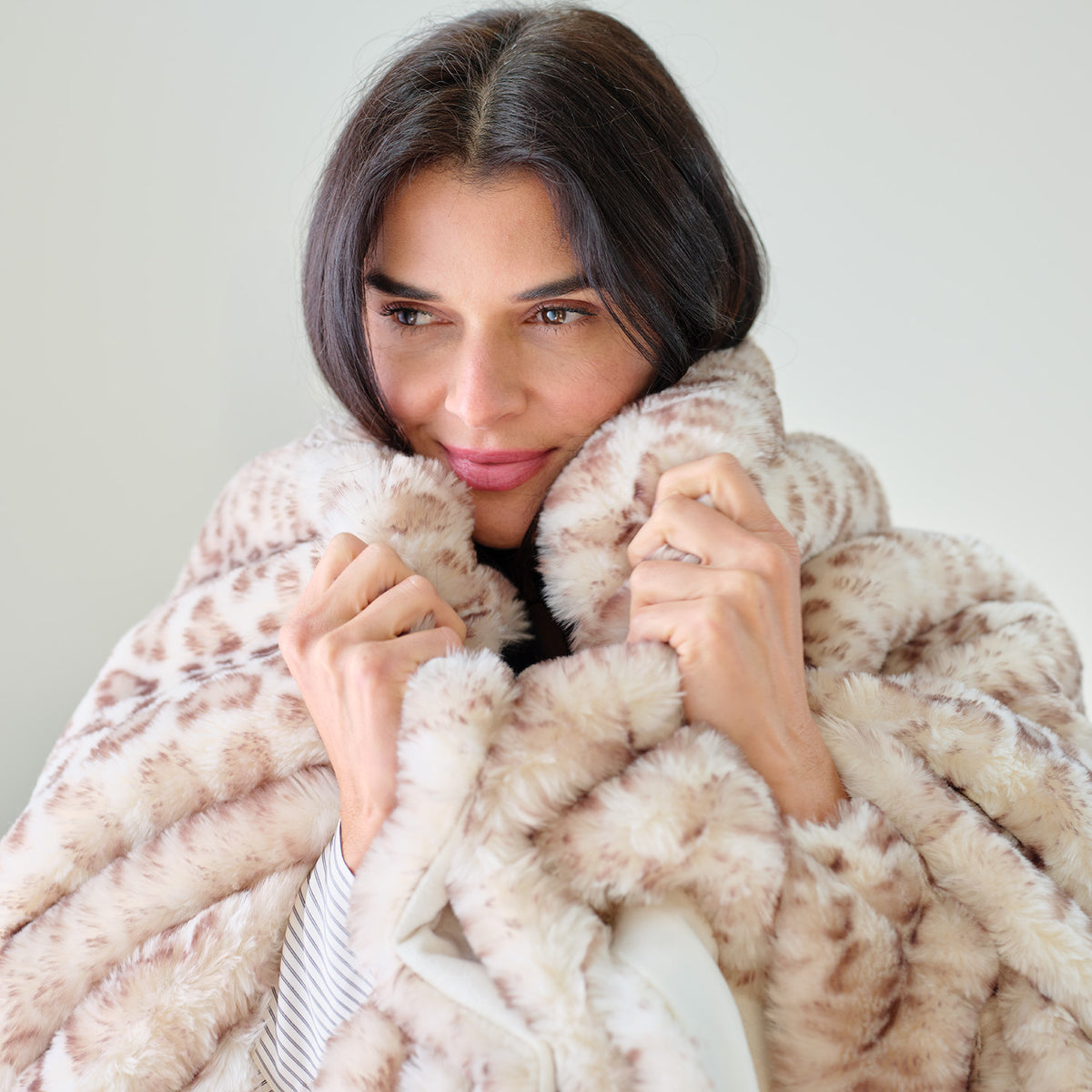 A woman with dark hair is wrapped in a plush faux fur throw blanket with a beige and brown leopard print pattern, holding the collar close to her face against a plain light background.