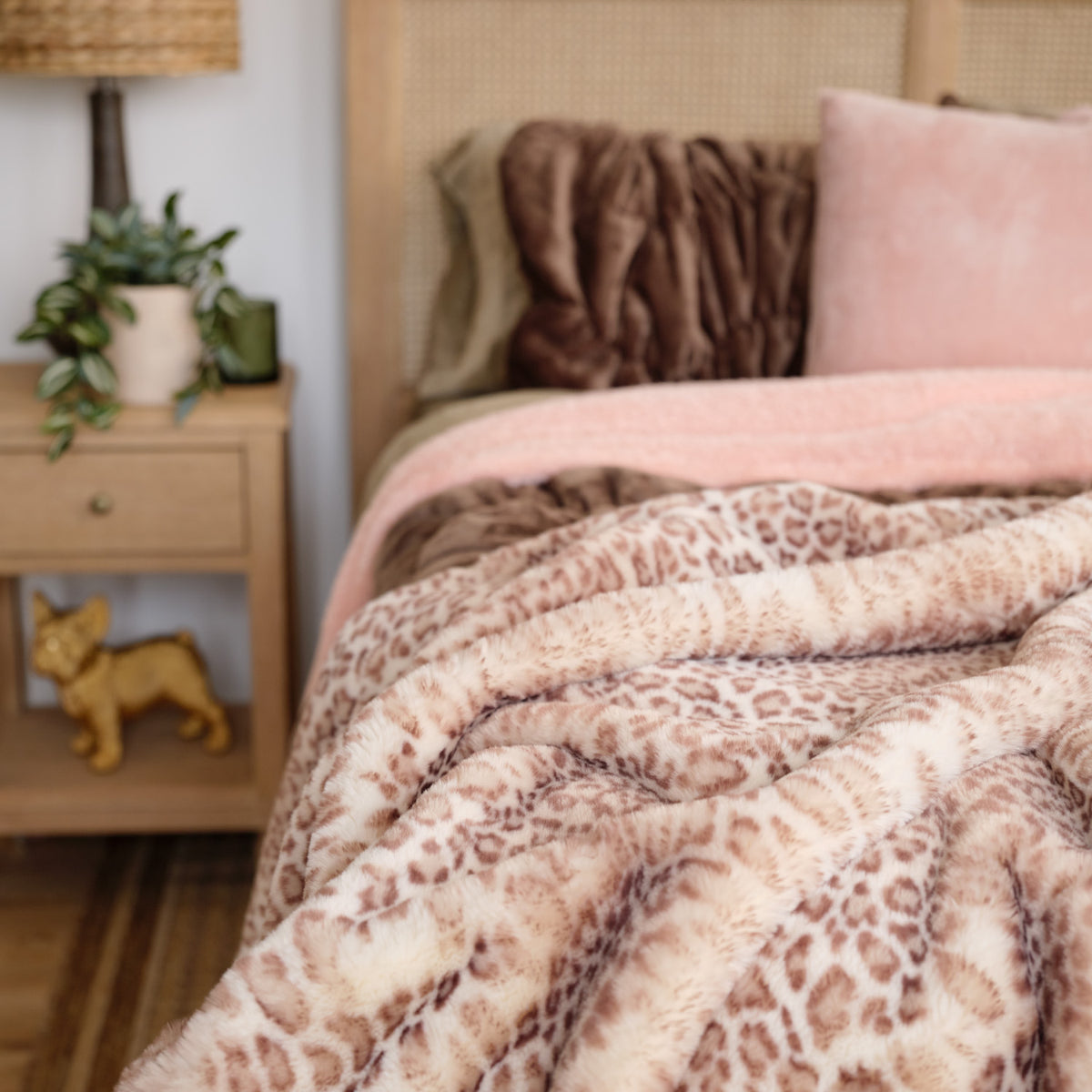 A plush leopard-print throw blanket in beige and brown is draped over a bed in a cozy bedroom setting, alongside a pink pillow and wooden bedside table.
