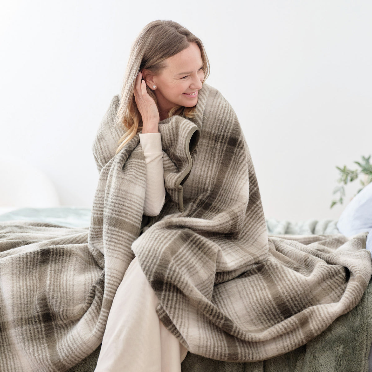 A woman sitting on a bed wrapped in a green plaid teddy fleece blanket.