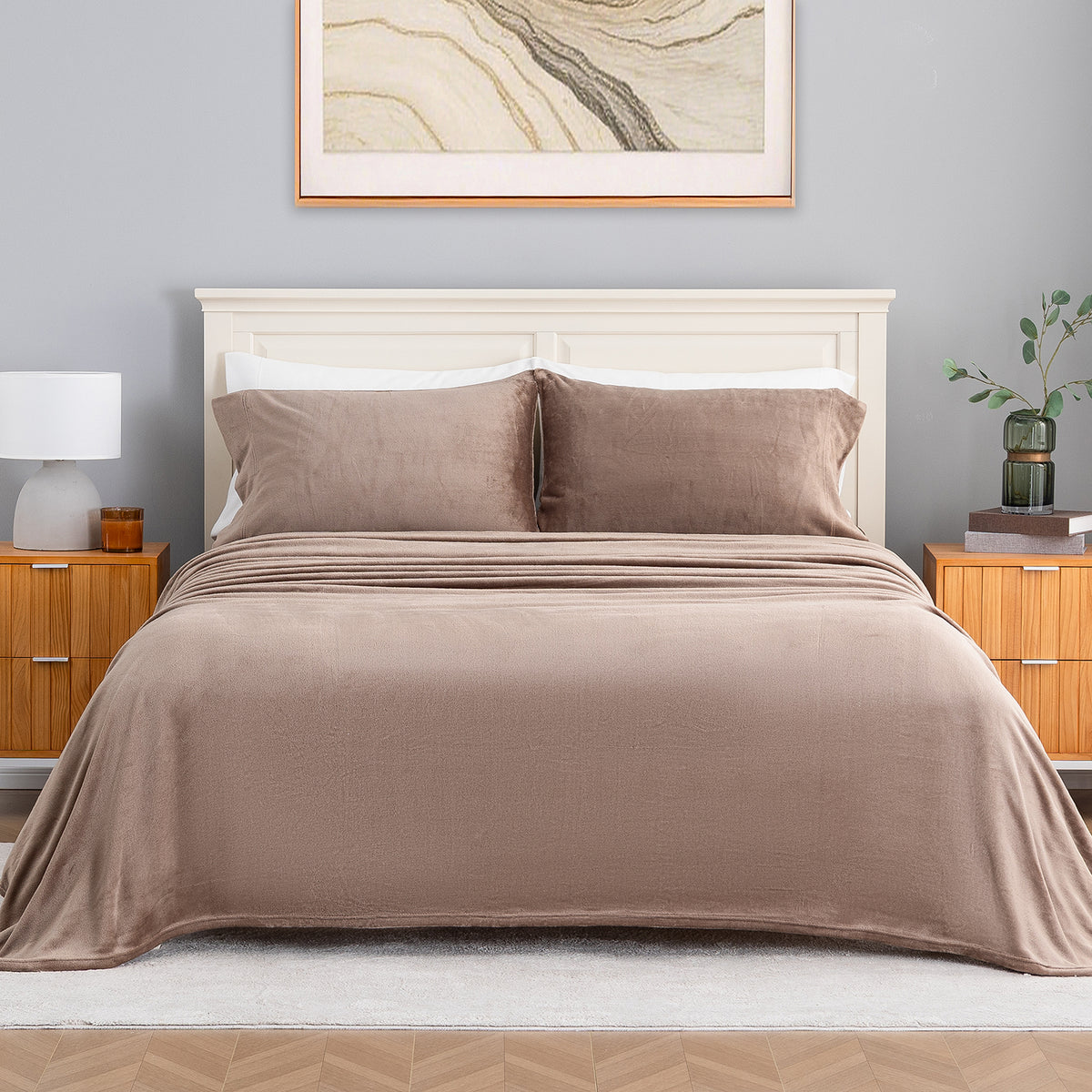 A neatly made bed with a smooth taupe-brown blanket and matching pillow shams, set in a styled bedroom with a white headboard, wooden nightstands, a gray wall, and framed wall art.