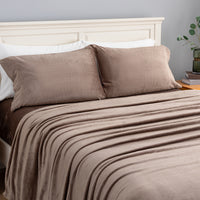 A neatly made bed with a taupe-brown blanket and two matching pillow shams, styled in a bedroom setting with a white wooden headboard, gray wall, and a small plant on a bedside table.