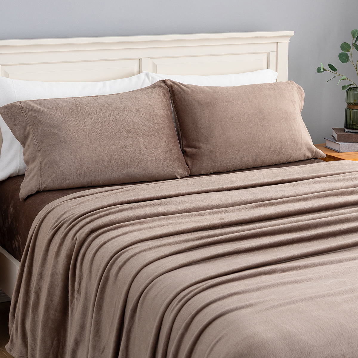 A neatly made bed with a taupe-brown blanket and two matching pillow shams, styled in a bedroom setting with a white wooden headboard, gray wall, and a small plant on a bedside table.