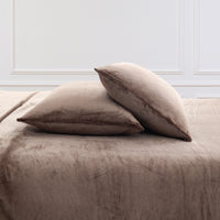 Two plush taupe throw pillows resting on a matching taupe bedspread against a white paneled wall.