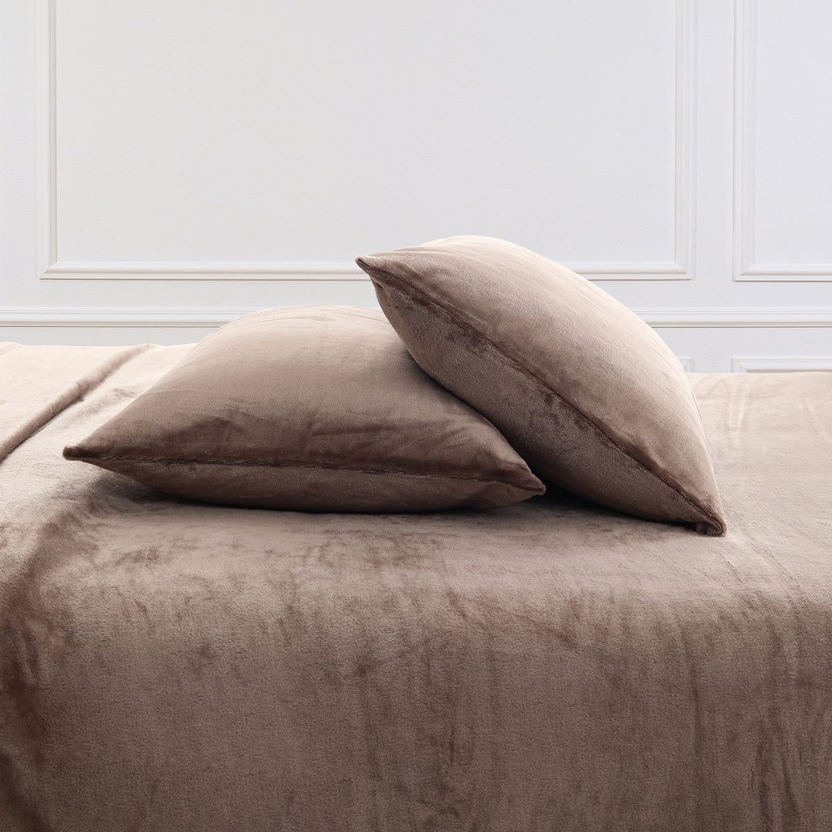 Two plush taupe throw pillows resting on a matching taupe bedspread against a white paneled wall.