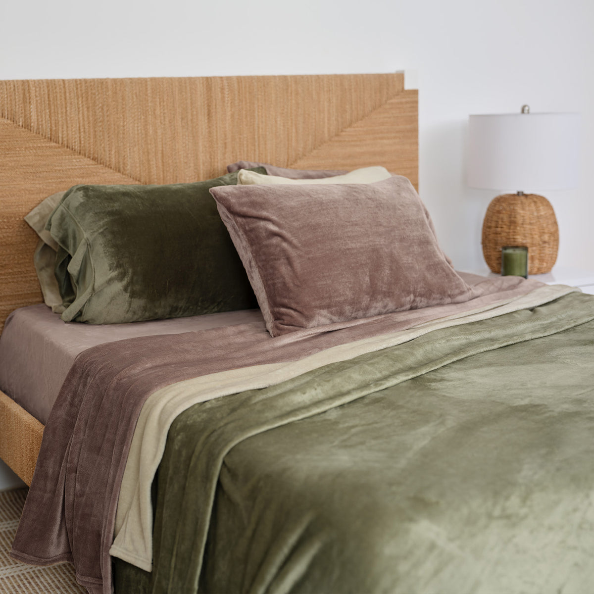 A styled bed with plush bedding in muted earthy tones, featuring an olive green throw blanket, a mauve velvet pillow, and a matching mauve throw, set against a light wood headboard and a white wall with a bedside lamp.