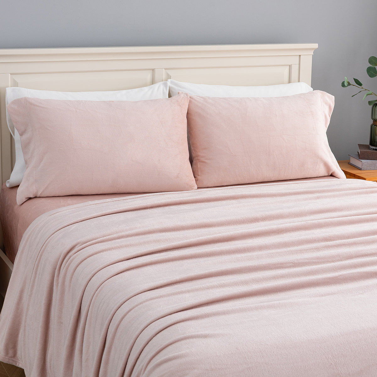 A neatly made bed with blush pink bedding, including two pillowcases and a matching blanket, styled with a cream-colored wooden headboard against a soft gray wall.